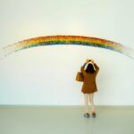 A woman stands observing a colorful rainbow art installation indoors.
